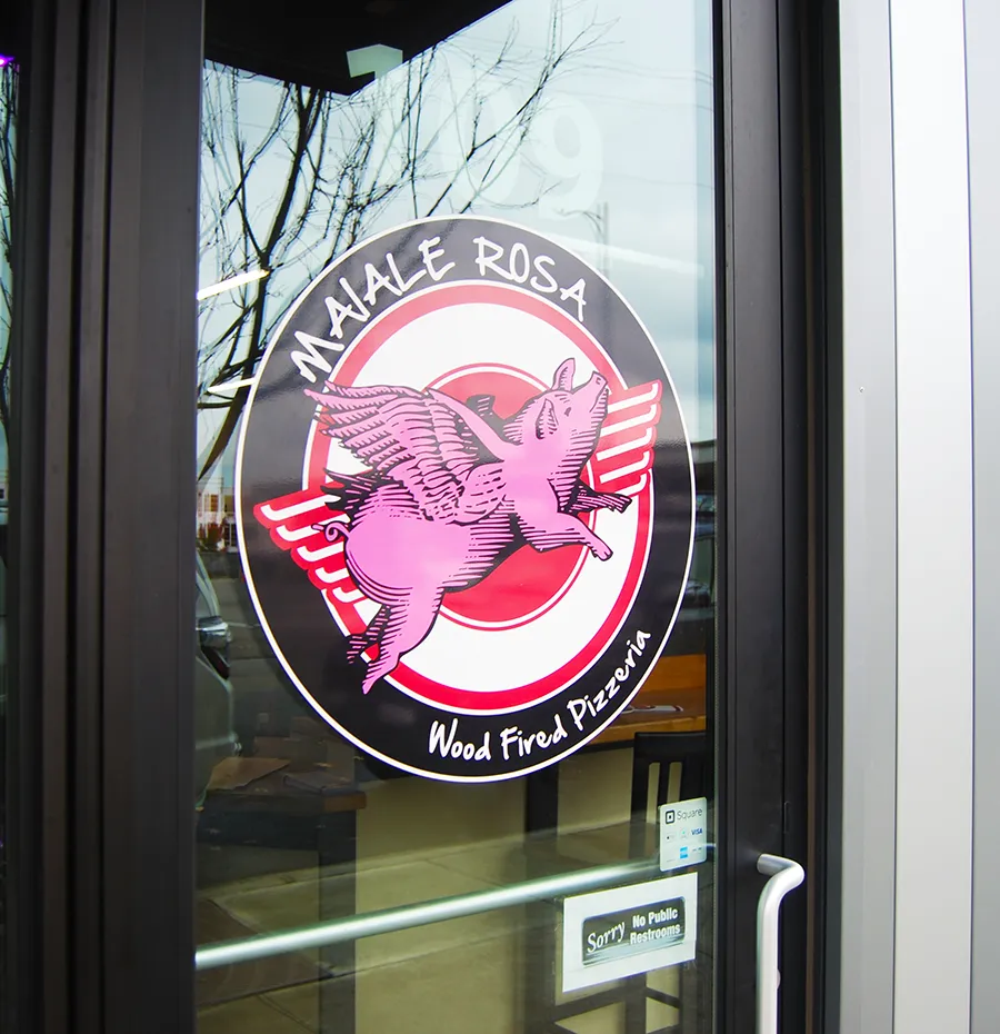 A glass door featuring the Maiale Rosa logo with the pink pig with wings on a red, white, and black background