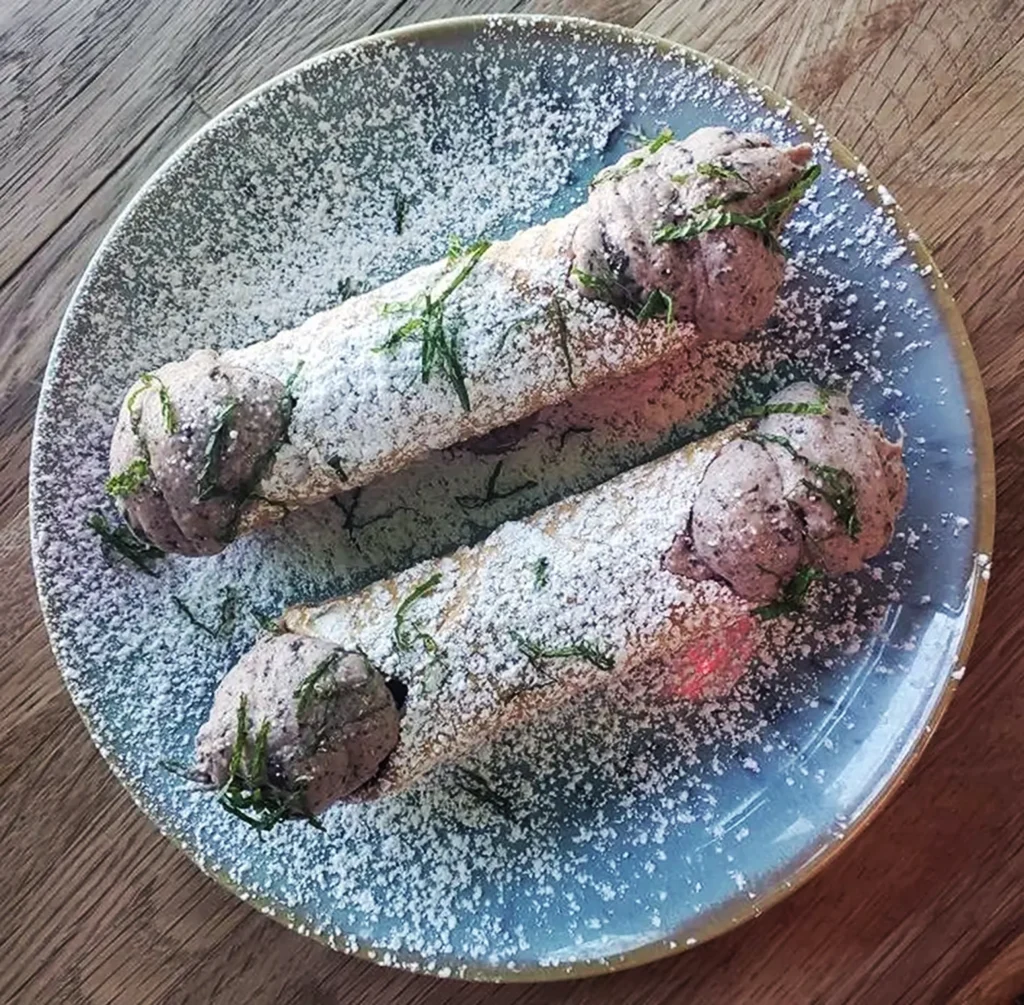 Two chocolate chip mint cannolis on a plate with powdered sugar on top.